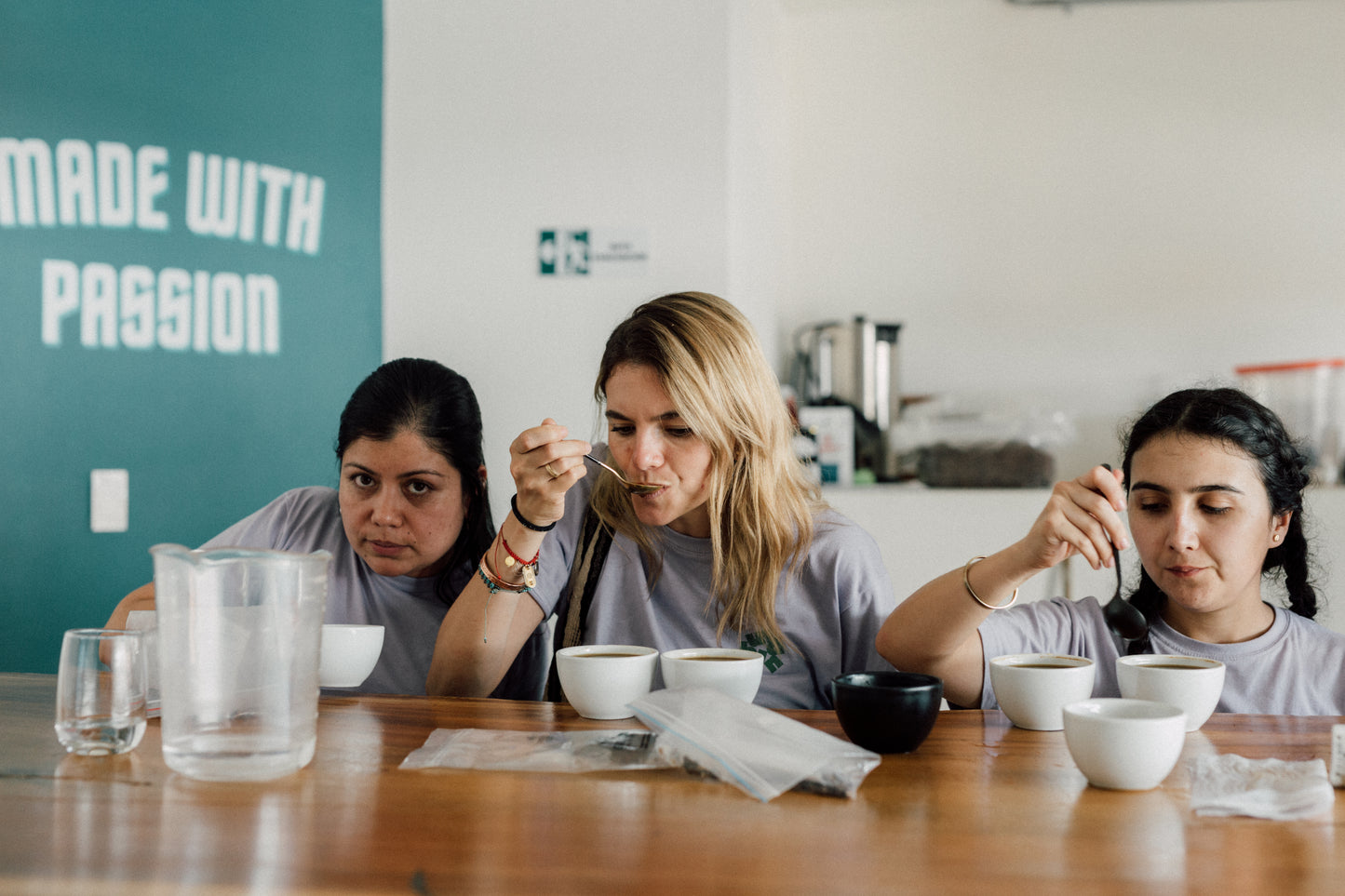 Three women tasting coffee at a table with a 'Made with Passion' sign in the background.