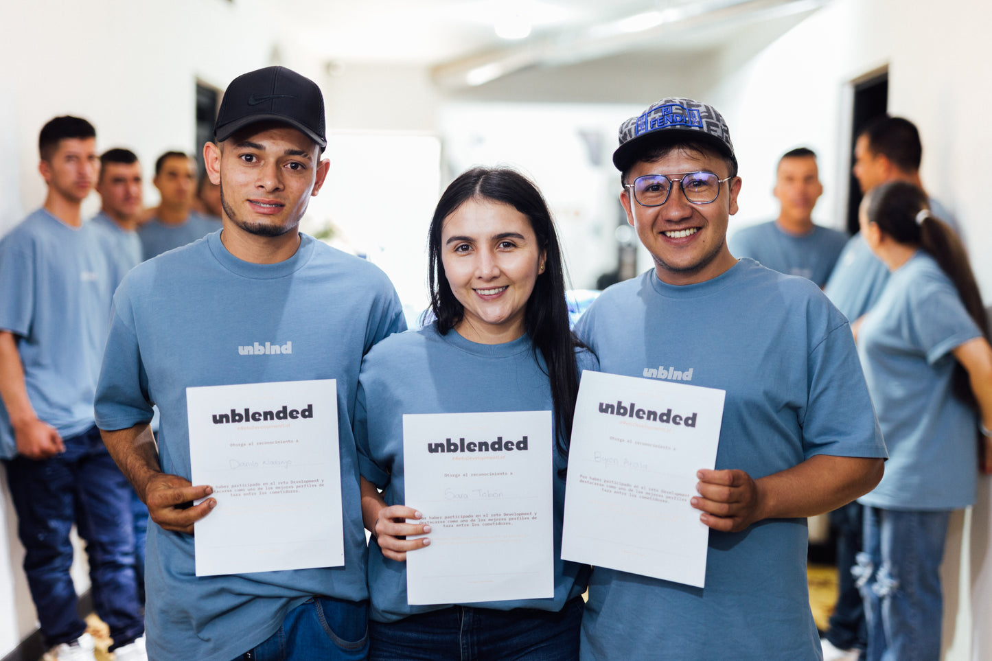 Three people wearing blue 'unblended' shirts holding certifications in a room with other people in the background.