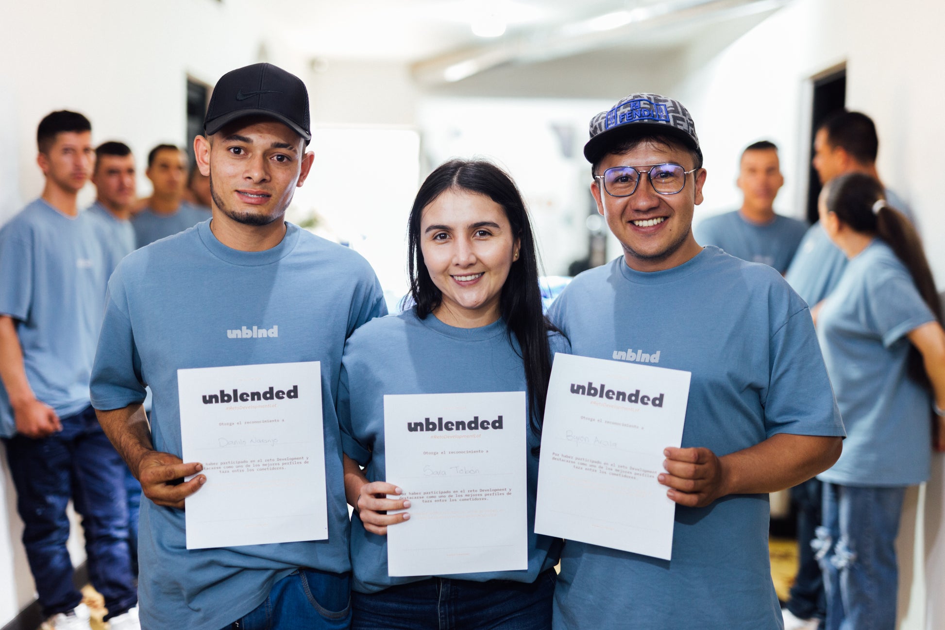 Three people wearing blue 'unblended' shirts holding certifications in a room with other people in the background.