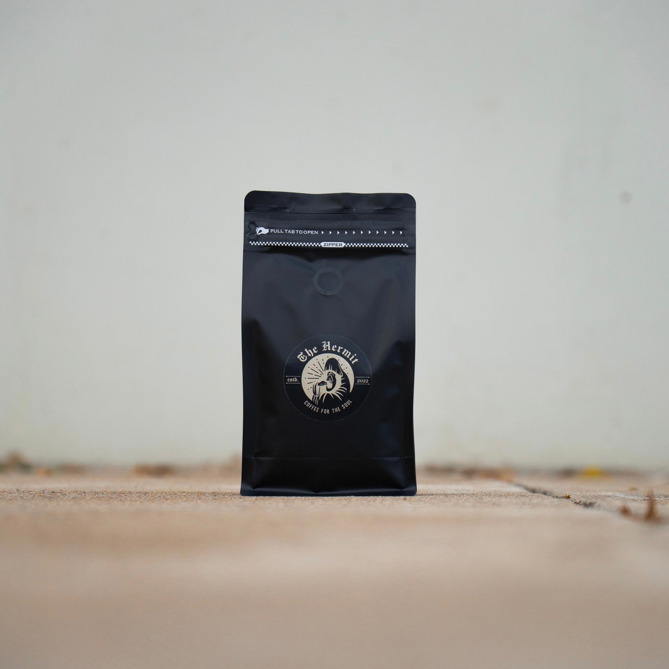 A black coffee bag with a gold and white logo, placed on a concrete surface with grass blades visible.