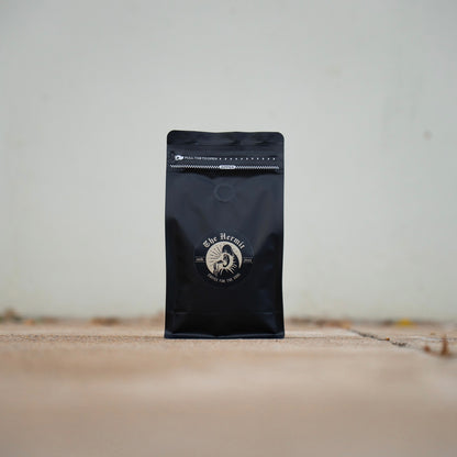 A black coffee bag with a gold and white logo, placed on a concrete surface with grass blades visible.