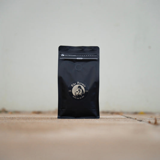 A black coffee bag with a gold and white logo, placed on a concrete surface with grass blades visible.